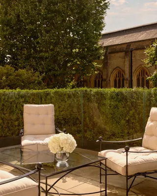 Private terrace with cushioned iron chairs and a glass table holding white hydrangeas, surrounded by greenery and views of a historic stone building with arched windows.