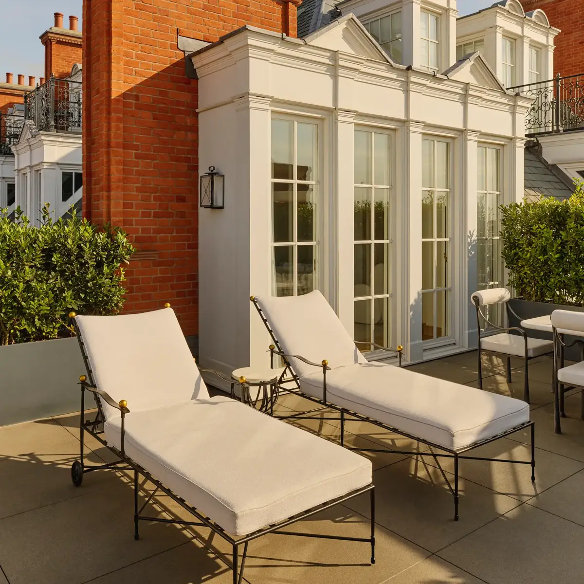Elegant terrace with two cushioned sun loungers and outdoor seating beside white French doors, set against red-brick architecture and trimmed greenery in warm afternoon light.