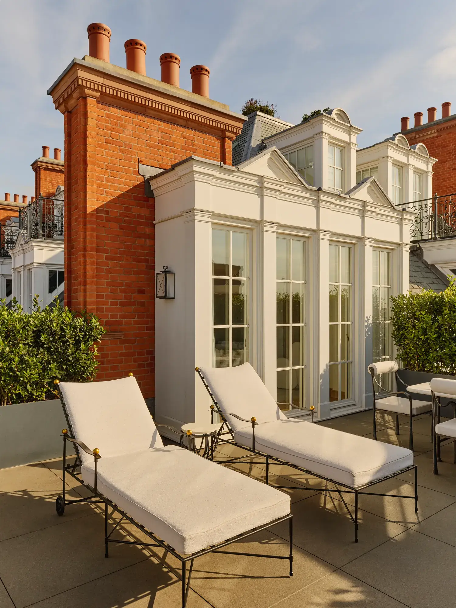 Elegant terrace with two cushioned sun loungers and outdoor seating beside white French doors, set against red-brick architecture and trimmed greenery in warm afternoon light.