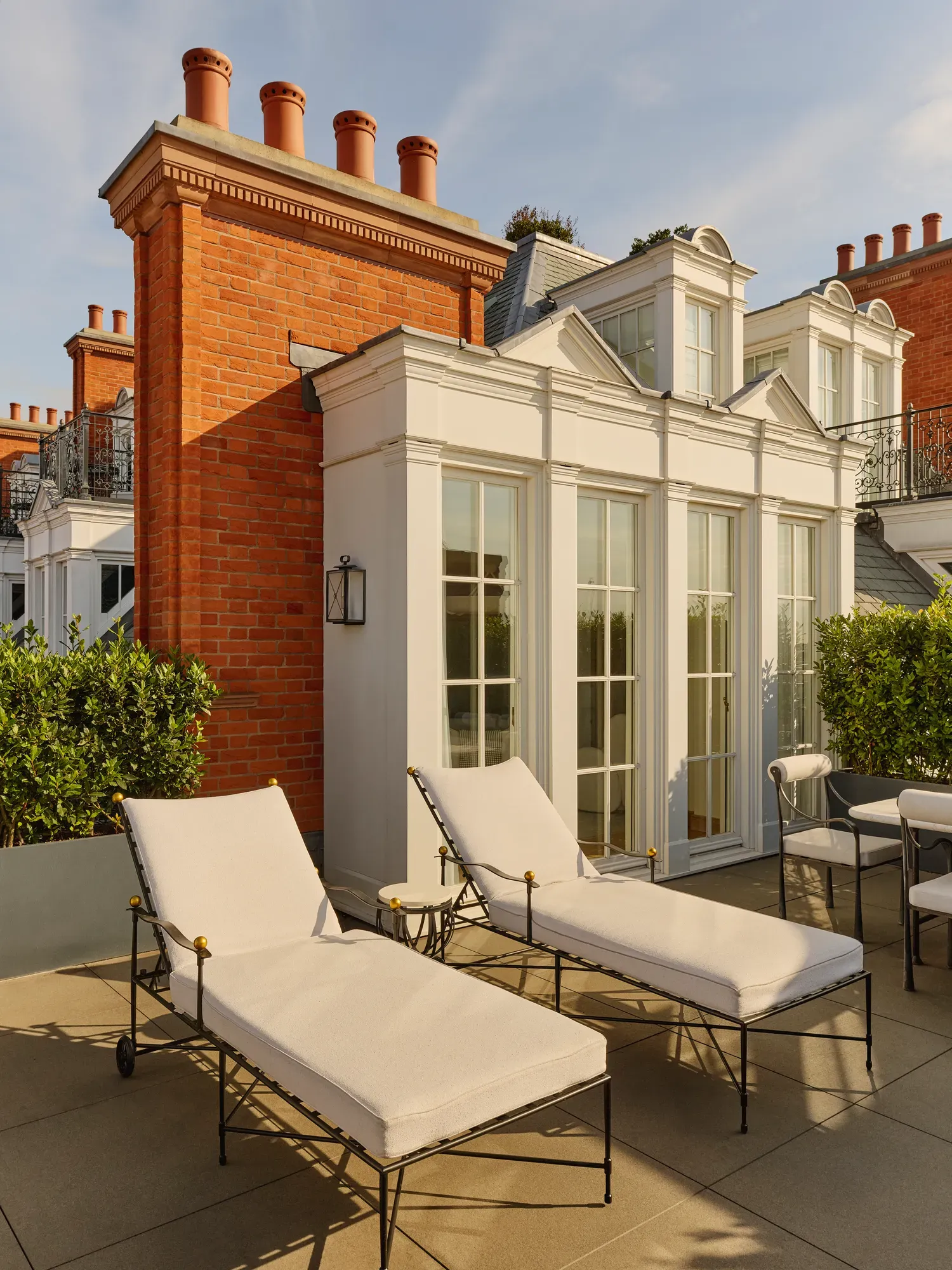 Elegant terrace with two cushioned sun loungers and outdoor seating beside white French doors, set against red-brick architecture and trimmed greenery in warm afternoon light.