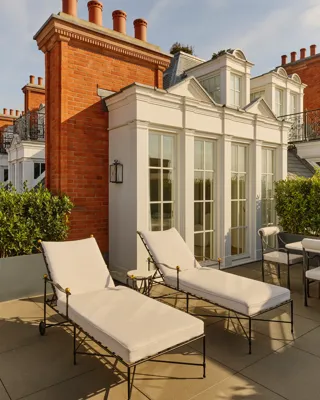 Elegant terrace with two cushioned sun loungers and outdoor seating beside white French doors, set against red-brick architecture and trimmed greenery in warm afternoon light.