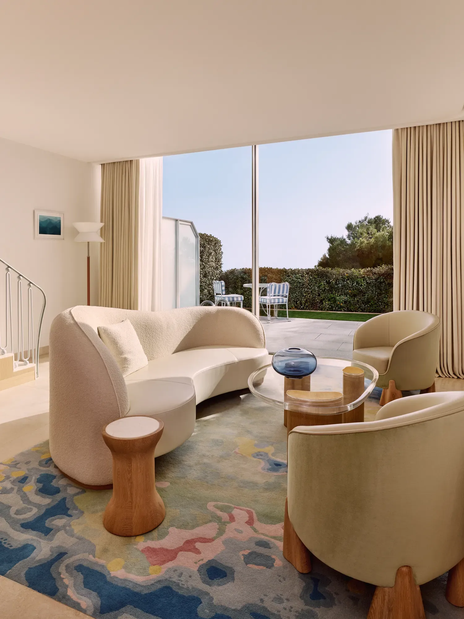 Bright living room with curved sofa, armchairs, and colourful rug, opening onto a terrace with garden views through floor-to-ceiling windows.