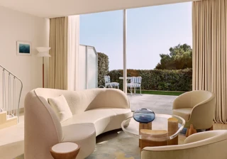 Bright living room with curved sofa, armchairs, and colourful rug, opening onto a terrace with garden views through floor-to-ceiling windows.