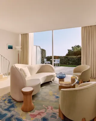Bright living room with curved sofa, armchairs, and colourful rug, opening onto a terrace with garden views through floor-to-ceiling windows.