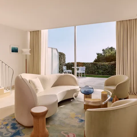 Salon lumineux avec canapé incurvé, fauteuils et tapis coloré, s’ouvrant sur une terrasse avec vue sur le jardin à travers de larges baies vitrées.