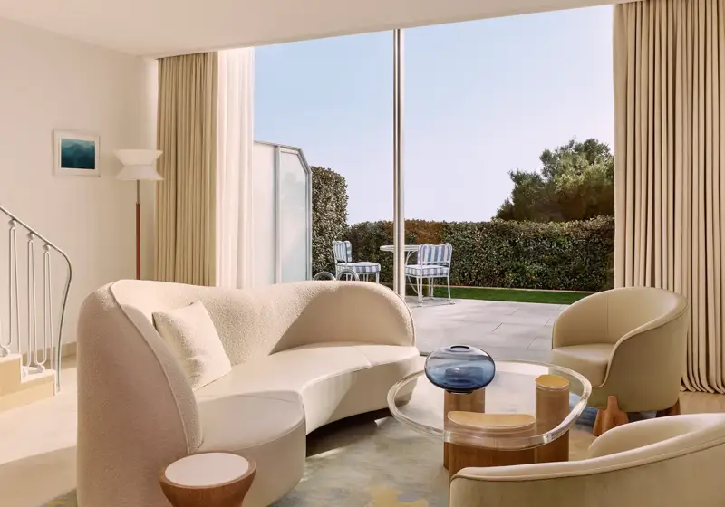 Bright living room with curved sofa, armchairs, and colourful rug, opening onto a terrace with garden views through floor-to-ceiling windows.