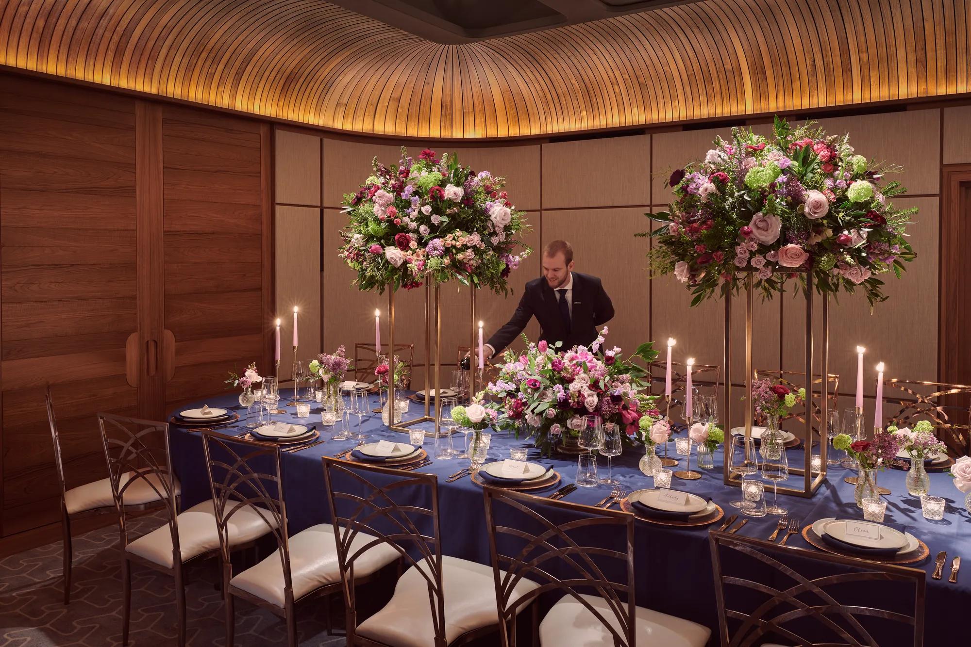 Private dining room with navy tablecloth, tall floral centerpieces, candles, and server pouring wine.