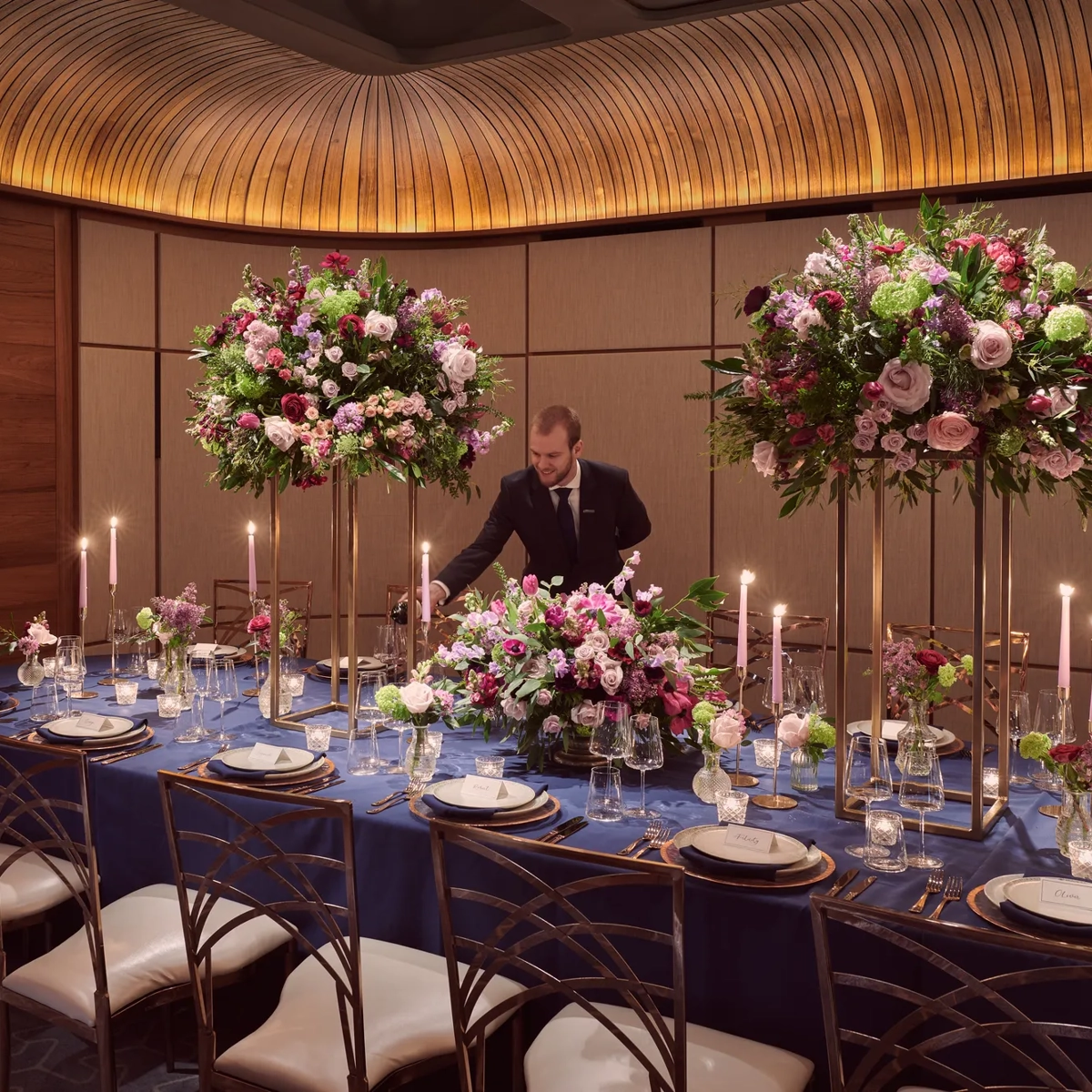Private dining room with navy tablecloth, tall floral centerpieces, candles, and server pouring wine.