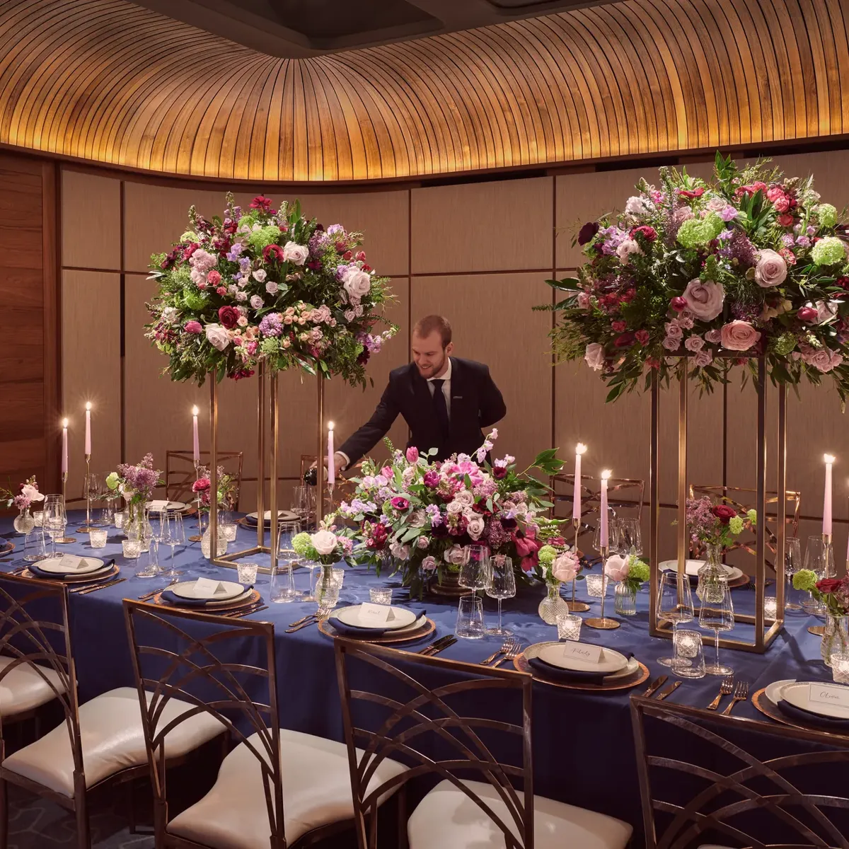 Private dining room with navy tablecloth, tall floral centerpieces, candles, and server pouring wine.