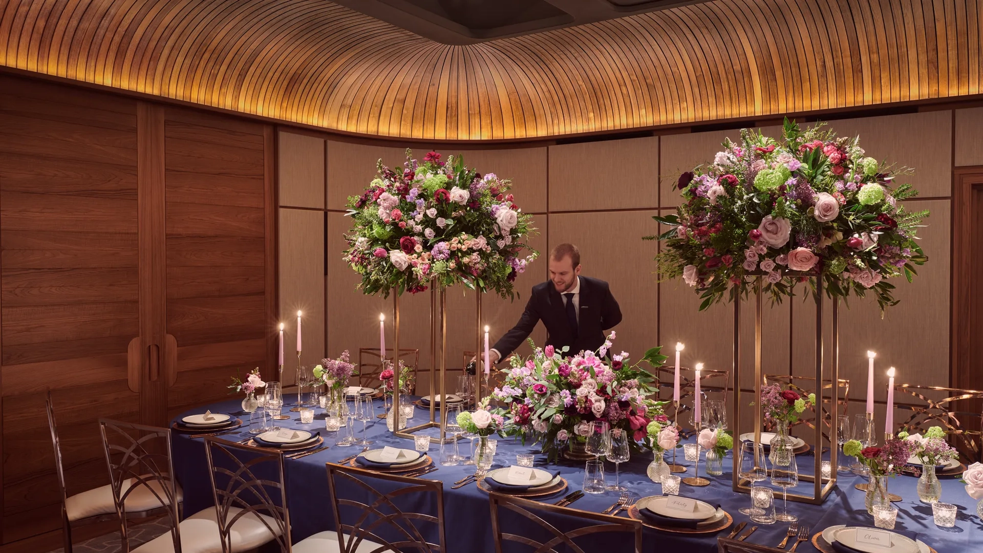 Private dining room with navy tablecloth, tall floral centerpieces, candles, and server pouring wine.