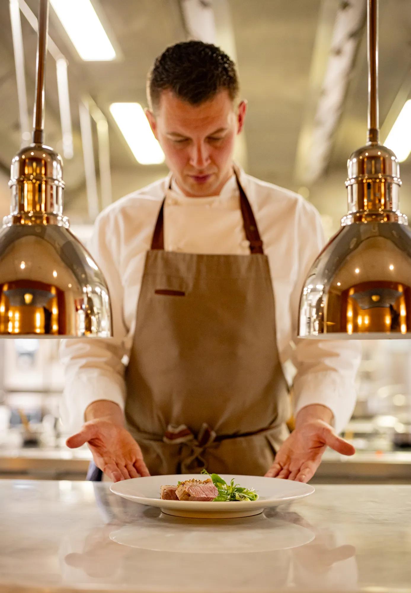 Chef presenting a plated dish under warm kitchen lights.