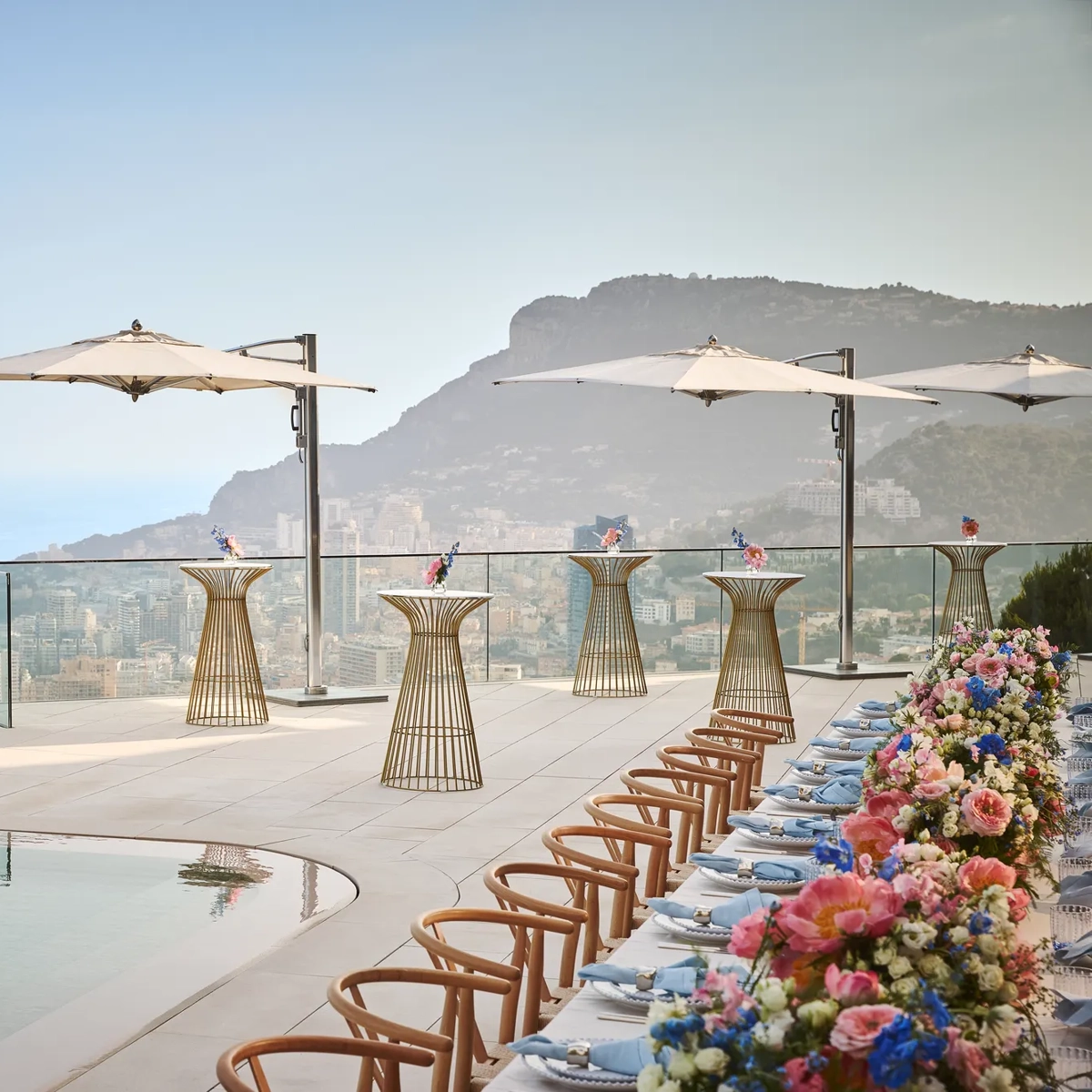 Terrasse sur le toit avec une table dressée au bord de la piscine, ornée de fleurs colorées et de chaises en bois, accompagnée de tables hautes avec parasols, dominant la ville et la mer en contrebas.
