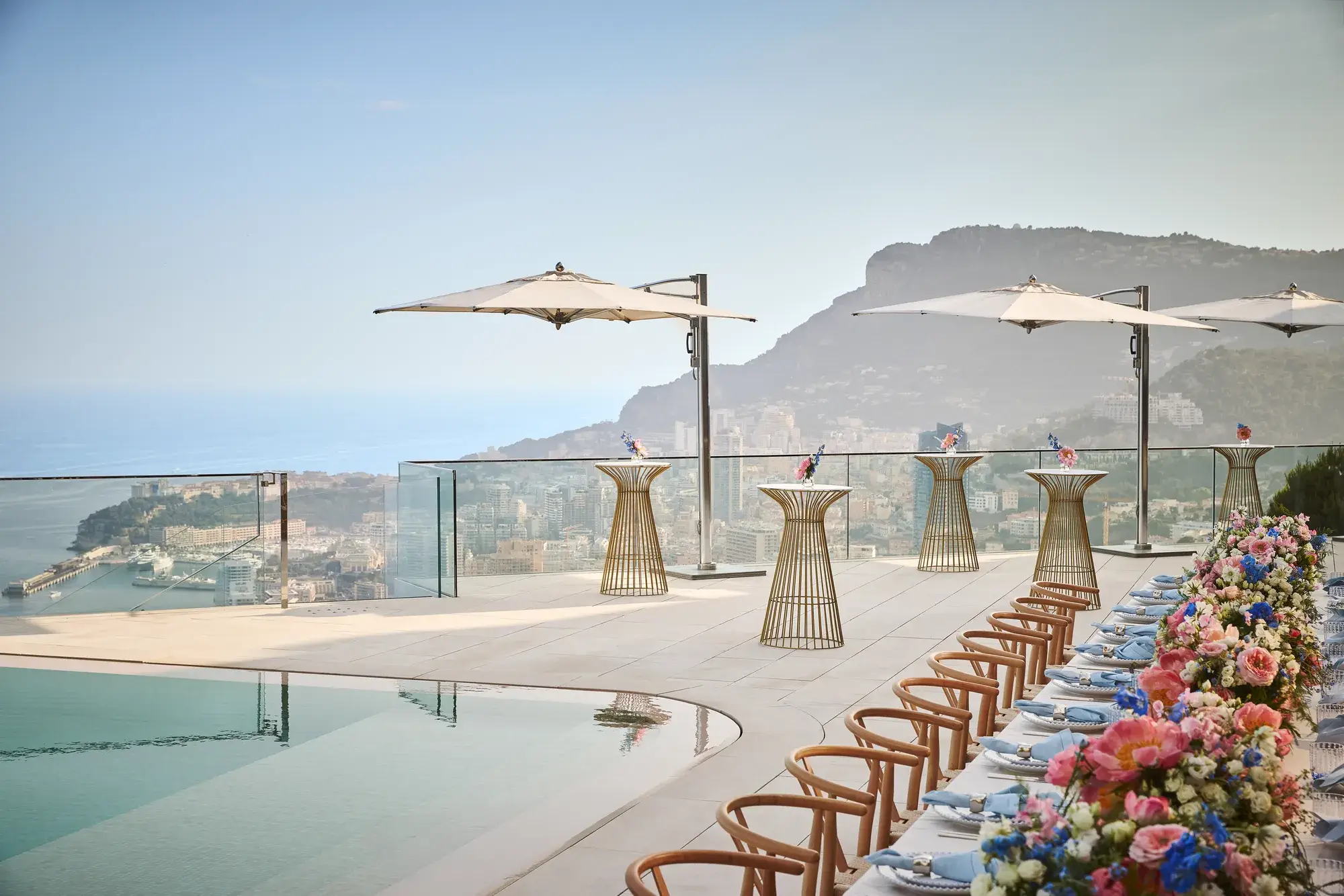 Terrasse sur le toit avec une table dressée au bord de la piscine, ornée de fleurs colorées et de chaises en bois, accompagnée de tables hautes avec parasols, dominant la ville et la mer en contrebas.