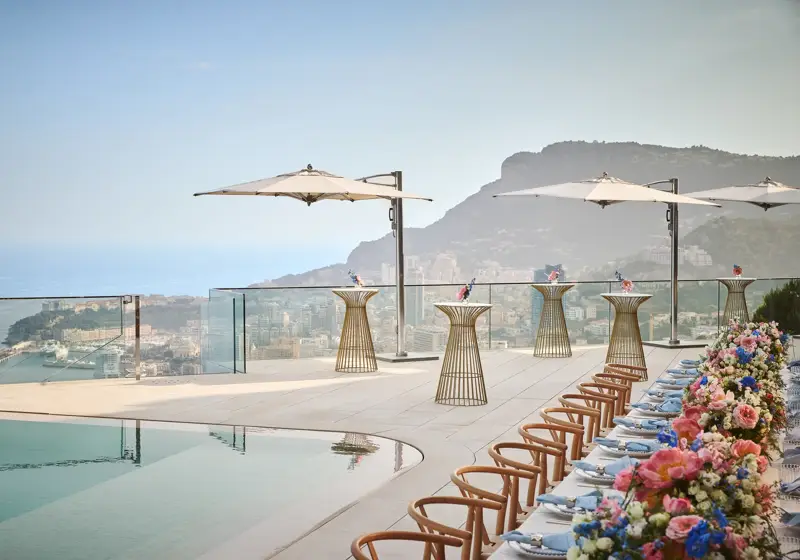 Terrasse sur le toit avec une table dressée au bord de la piscine, ornée de fleurs colorées et de chaises en bois, accompagnée de tables hautes avec parasols, dominant la ville et la mer en contrebas.