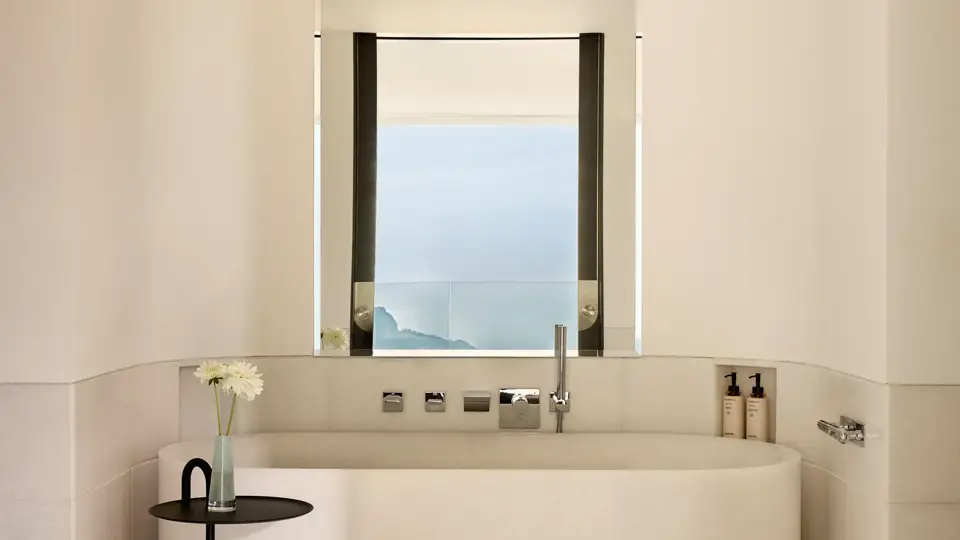 Minimalist bathroom with freestanding bathtub beneath a window, offering sea views, accompanied by a small side table with a single flower.