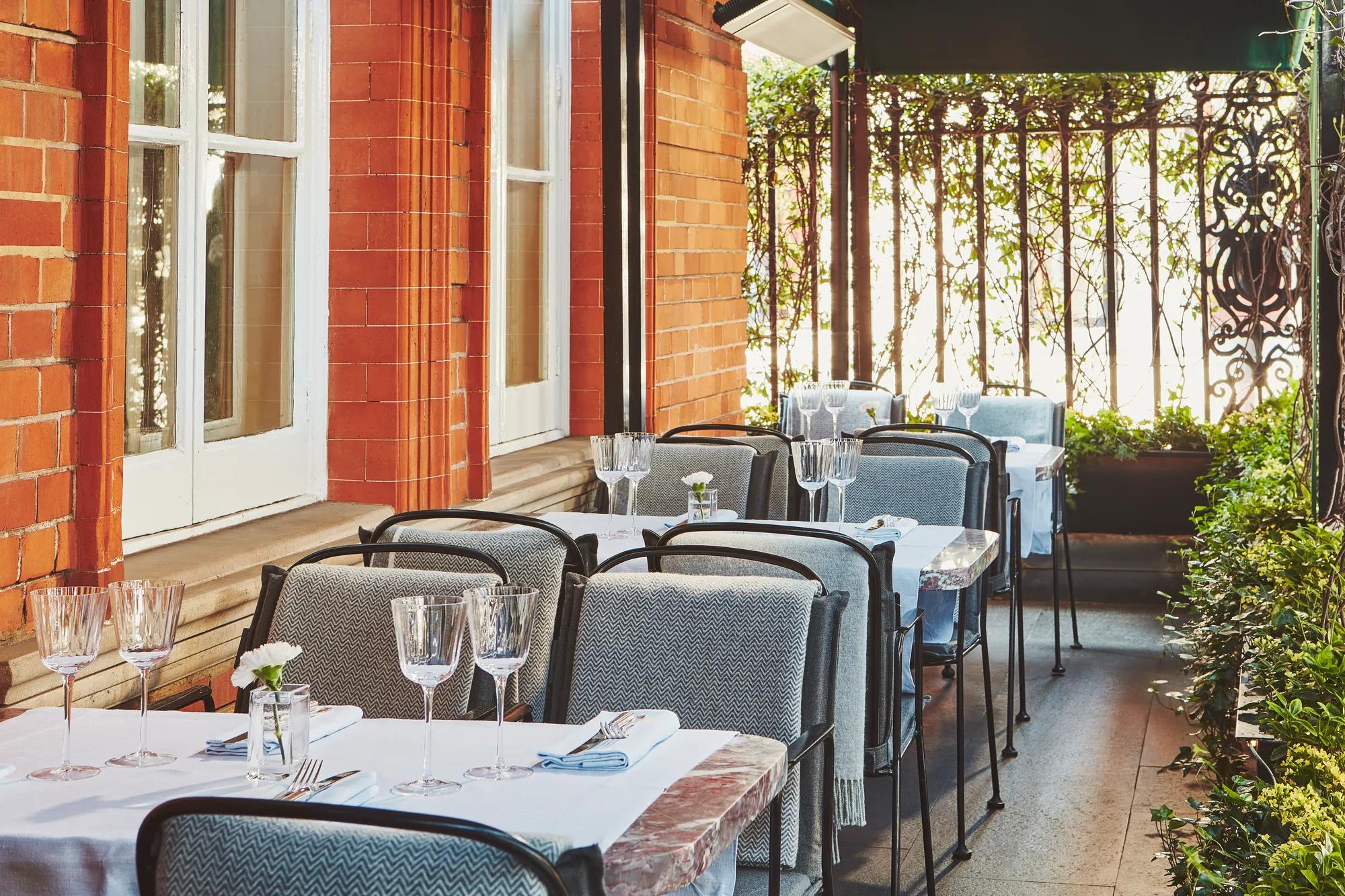 Outdoor terrace with white-clothed tables, glassware, and cushioned chairs beside a red brick wall and wrought-iron railing.