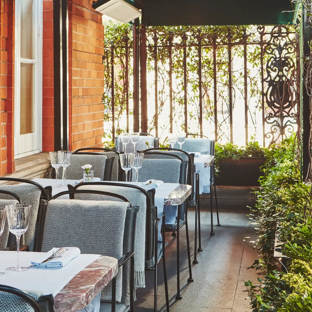Outdoor terrace with white-clothed tables, glassware, and cushioned chairs beside a red brick wall and wrought-iron railing.