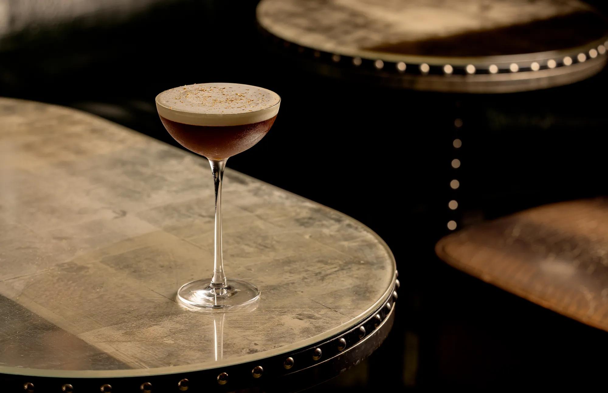 Dark cocktail with foam top in coupe glass on studded marble table.