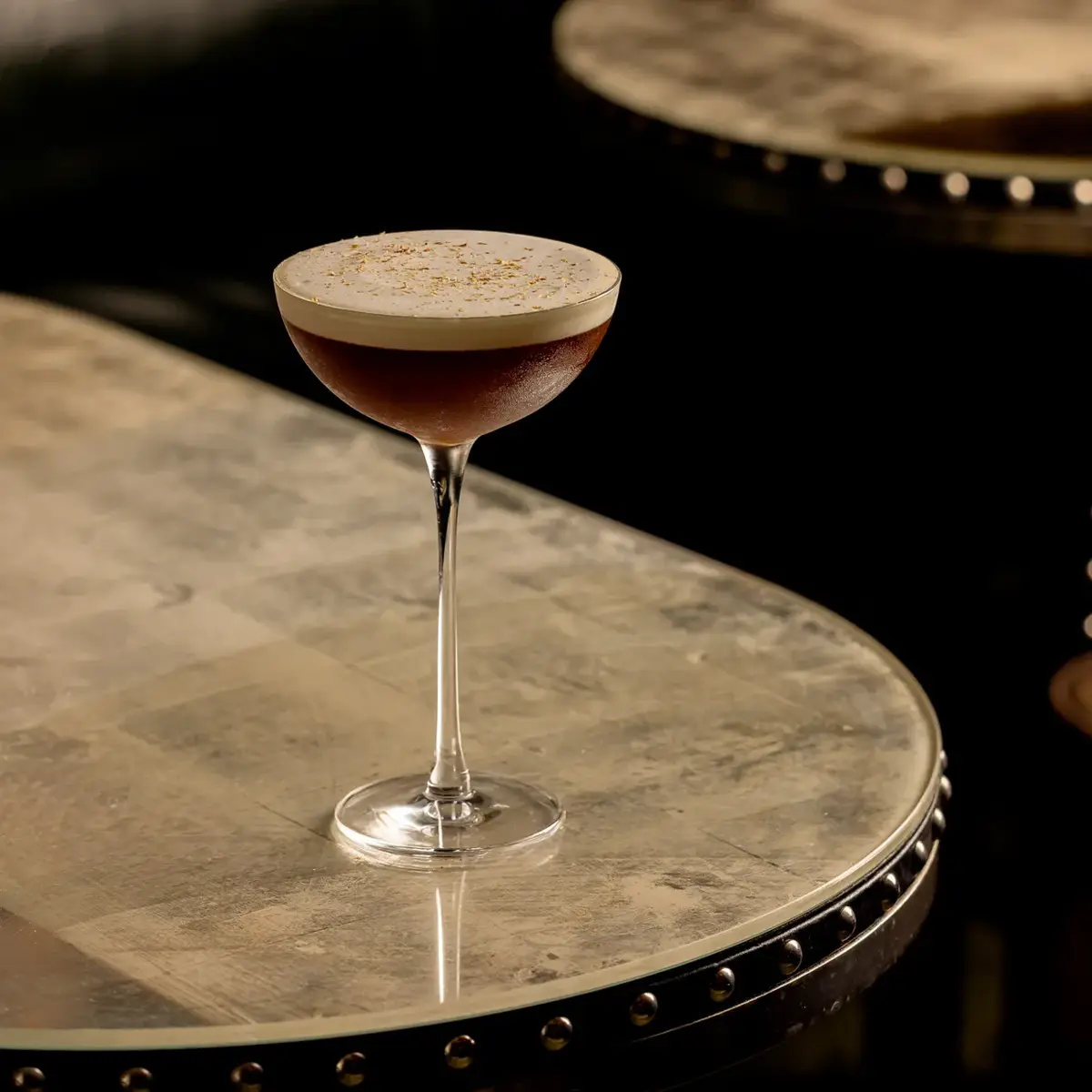 Dark cocktail with foam top in coupe glass on studded marble table.