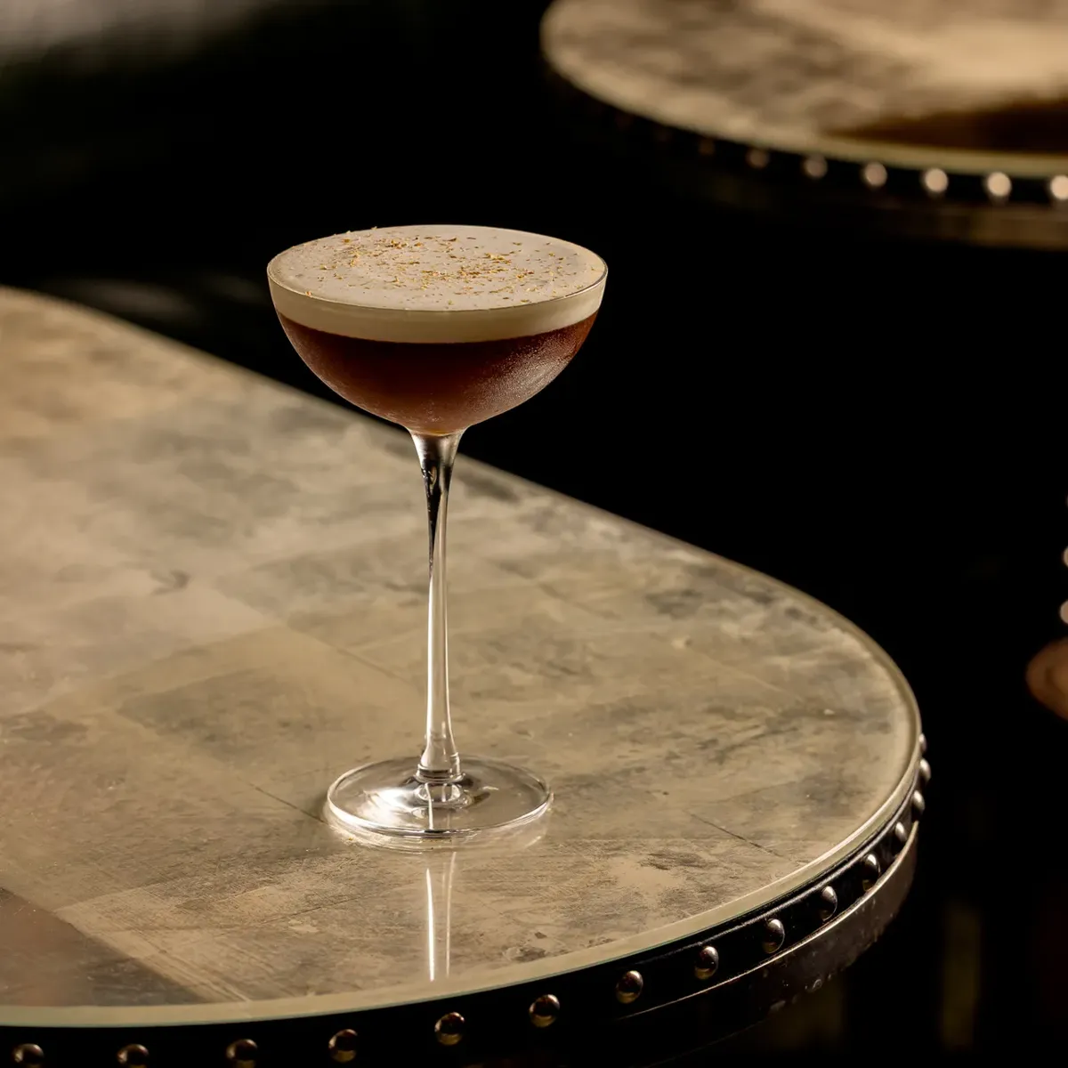 Dark cocktail with foam top in coupe glass on studded marble table.