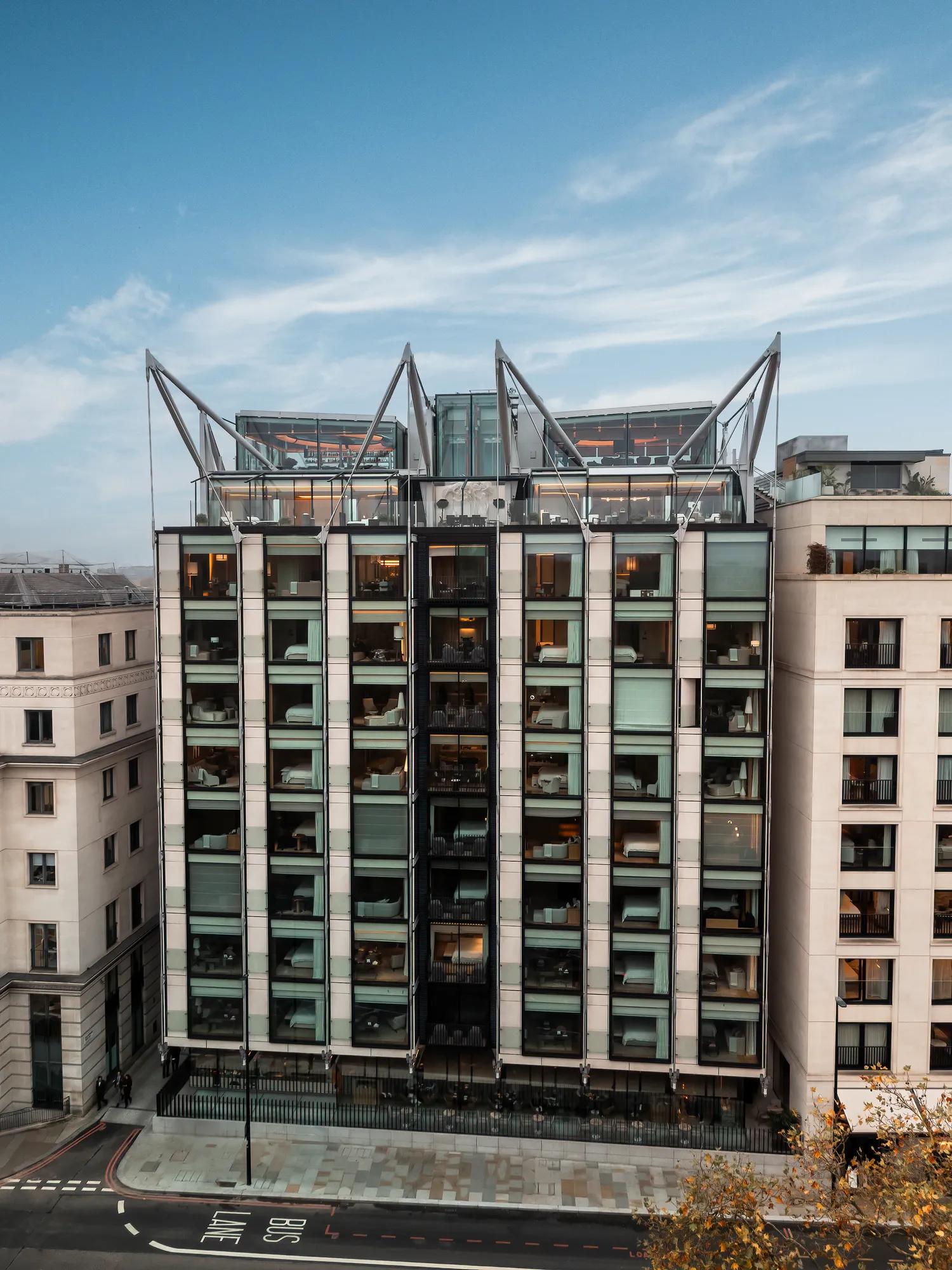 Modern glass-fronted hotel building with balconies and striking steel rooftop frames, viewed from across the street under a clear blue sky.