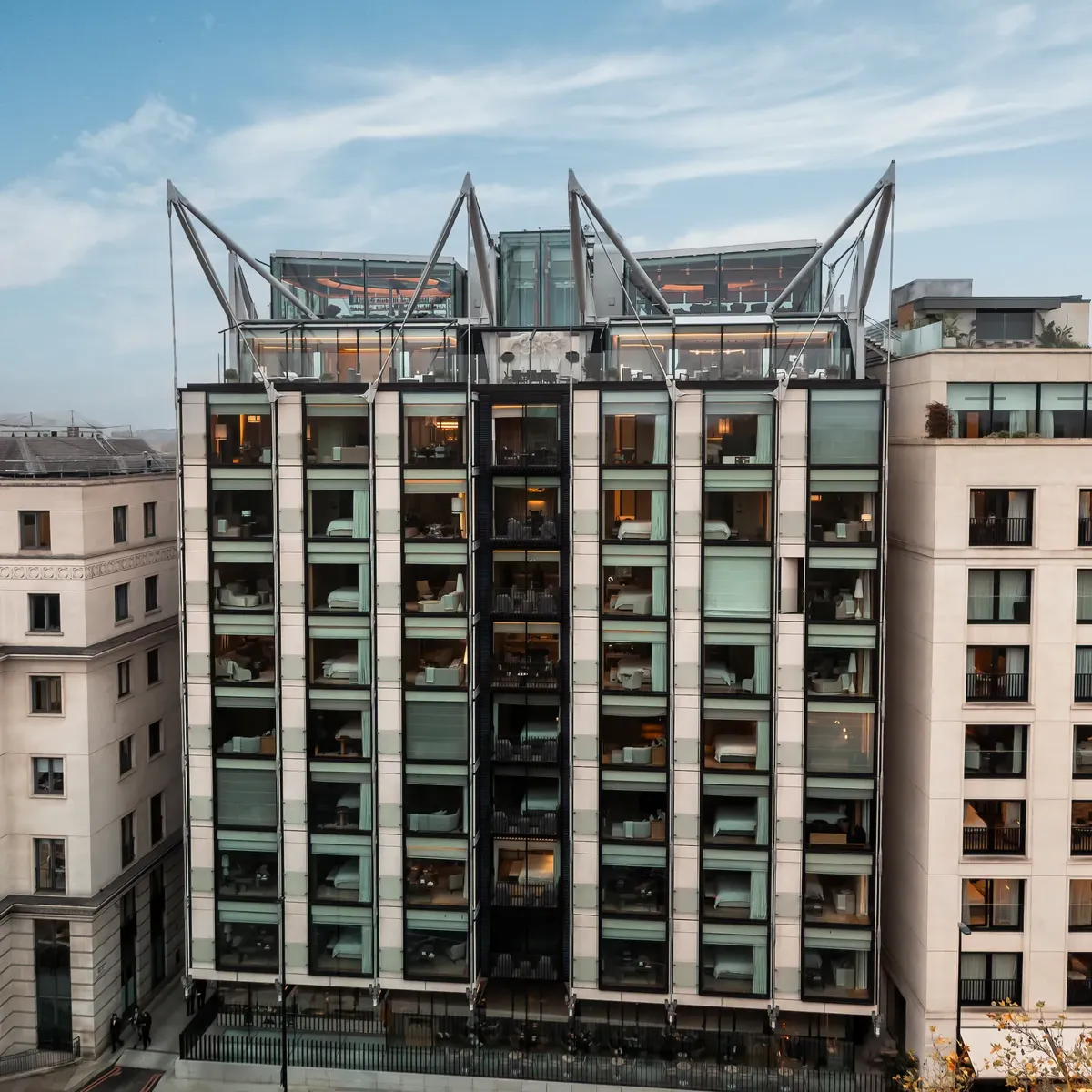 Modern glass-fronted hotel building with balconies and striking steel rooftop frames, viewed from across the street under a clear blue sky.