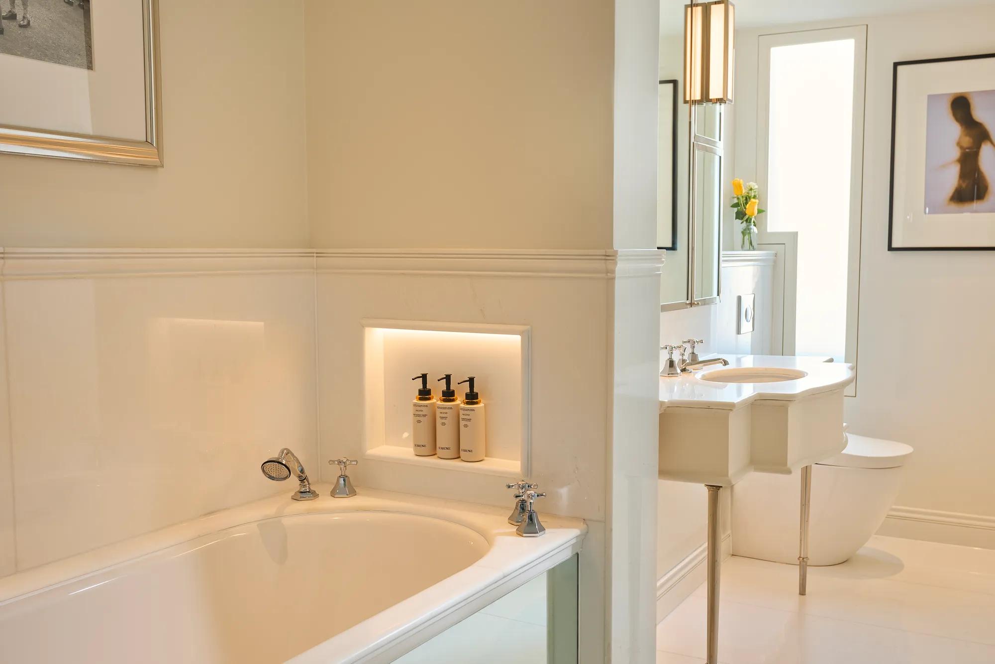 Bright marble bathroom with bathtub, vanity sink, and toilet in adjoining alcove.