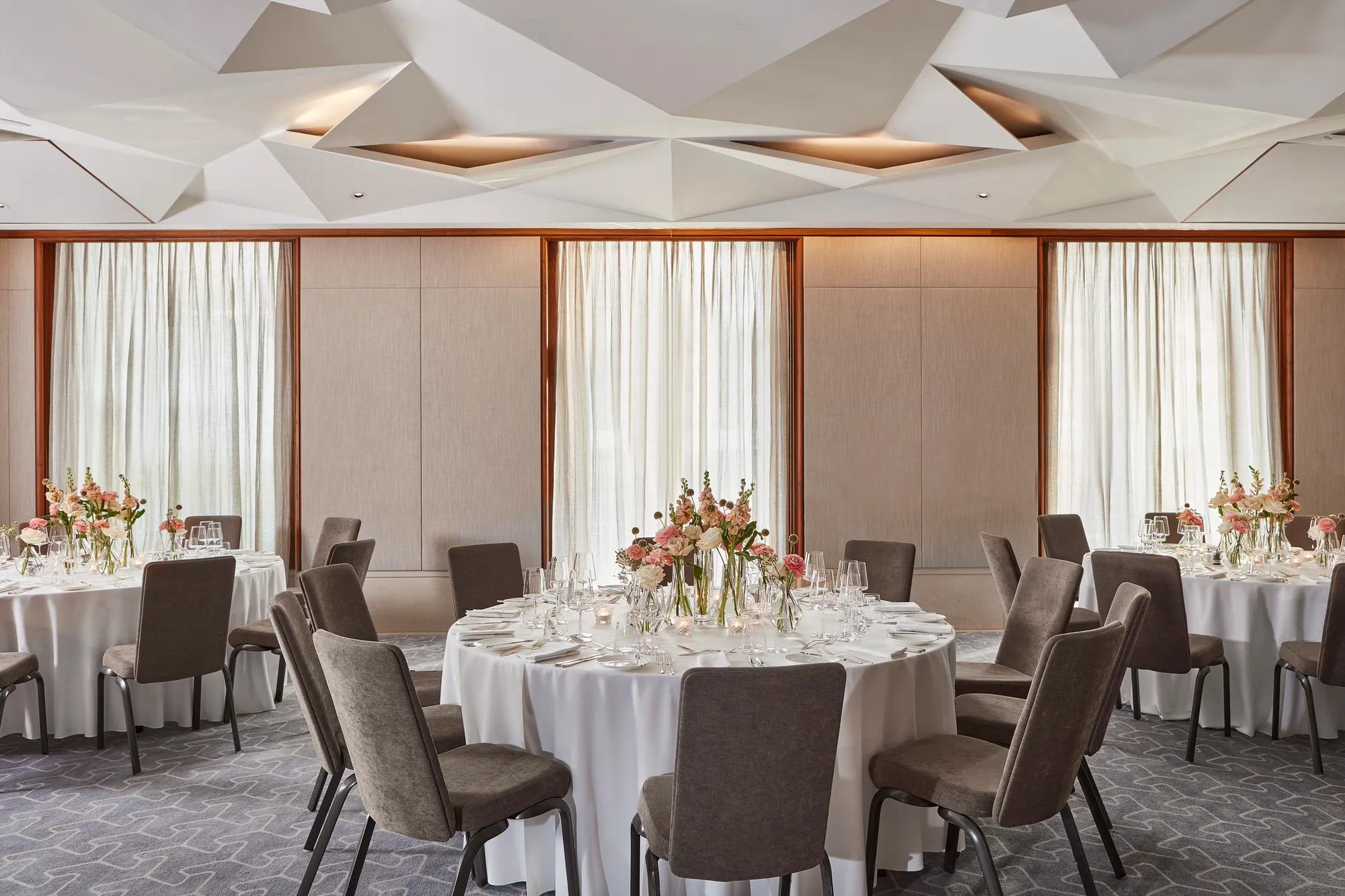 Elegant dining room with round tables, white tablecloths, floral centerpieces, and natural light through tall windows.