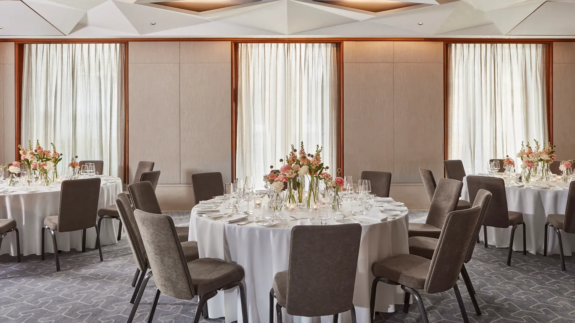 Elegant dining room with round tables, white tablecloths, floral centerpieces, and natural light through tall windows.