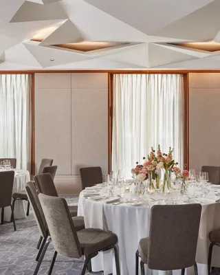 Elegant dining room with round tables, white tablecloths, floral centerpieces, and natural light through tall windows.