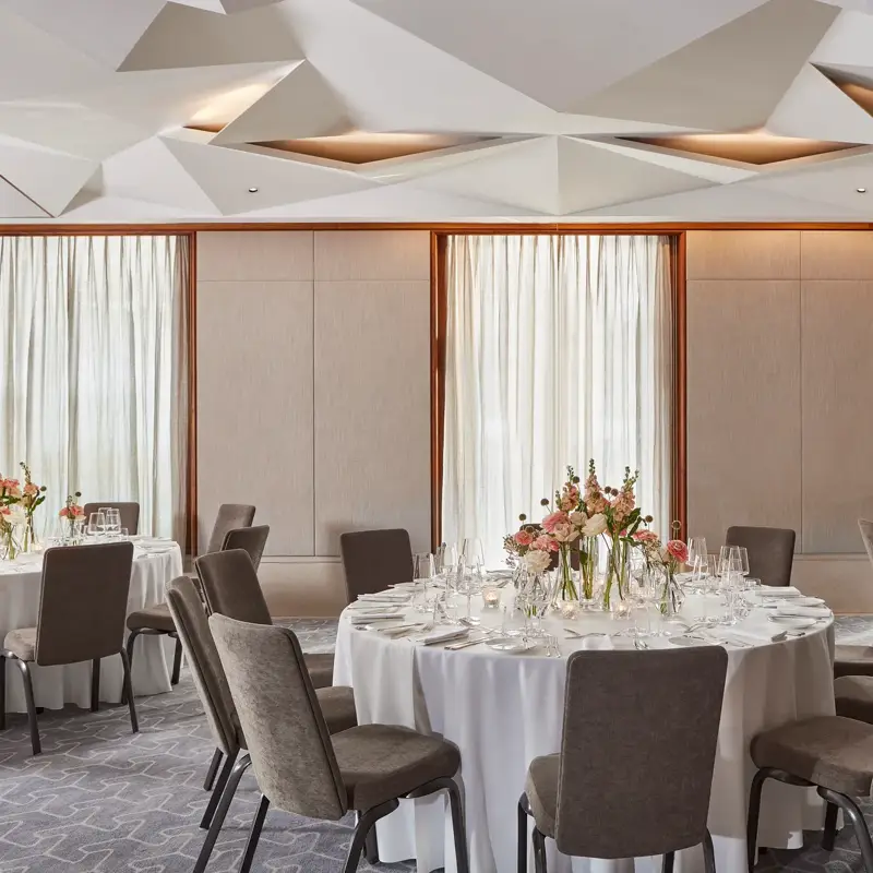 Elegant dining room with round tables, white tablecloths, floral centerpieces, and natural light through tall windows.