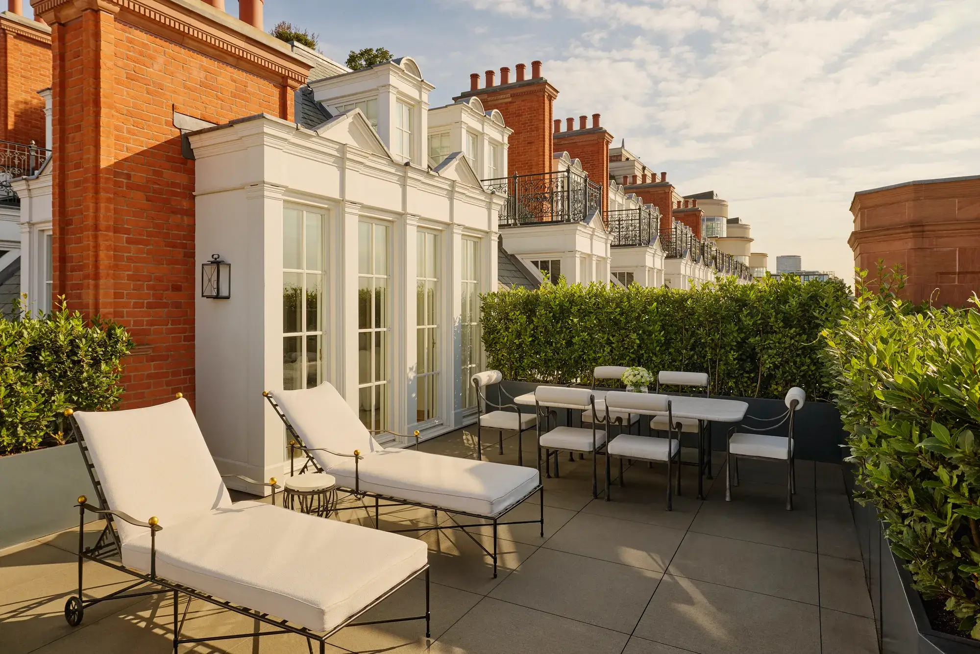 Private rooftop terrace with cushioned sun loungers and an outdoor dining table surrounded by greenery, set against red brick façades and white French doors in soft afternoon light.