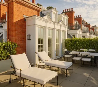 Private rooftop terrace with cushioned sun loungers and an outdoor dining table surrounded by greenery, set against red brick façades and white French doors in soft afternoon light.