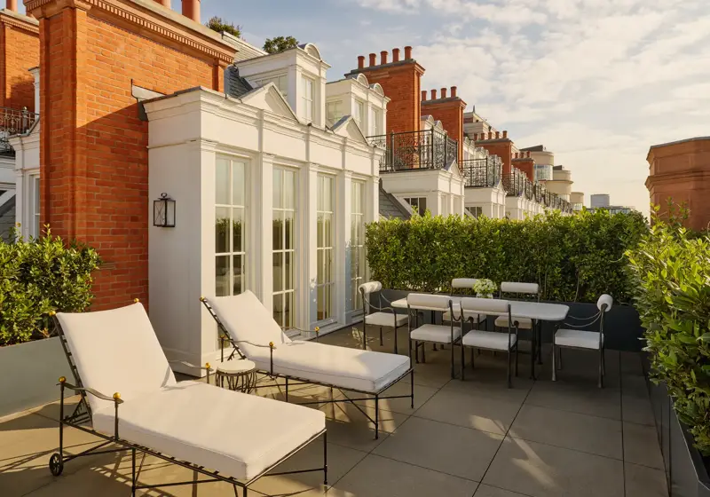 Private rooftop terrace with cushioned sun loungers and an outdoor dining table surrounded by greenery, set against red brick façades and white French doors in soft afternoon light.