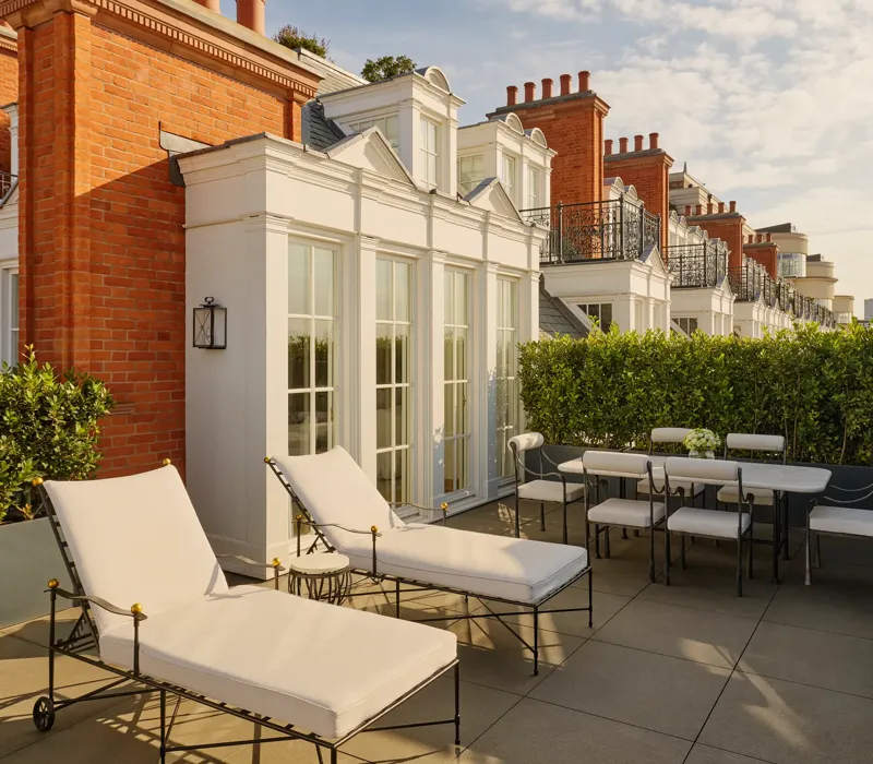 Private rooftop terrace with cushioned sun loungers and an outdoor dining table surrounded by greenery, set against red brick façades and white French doors in soft afternoon light.