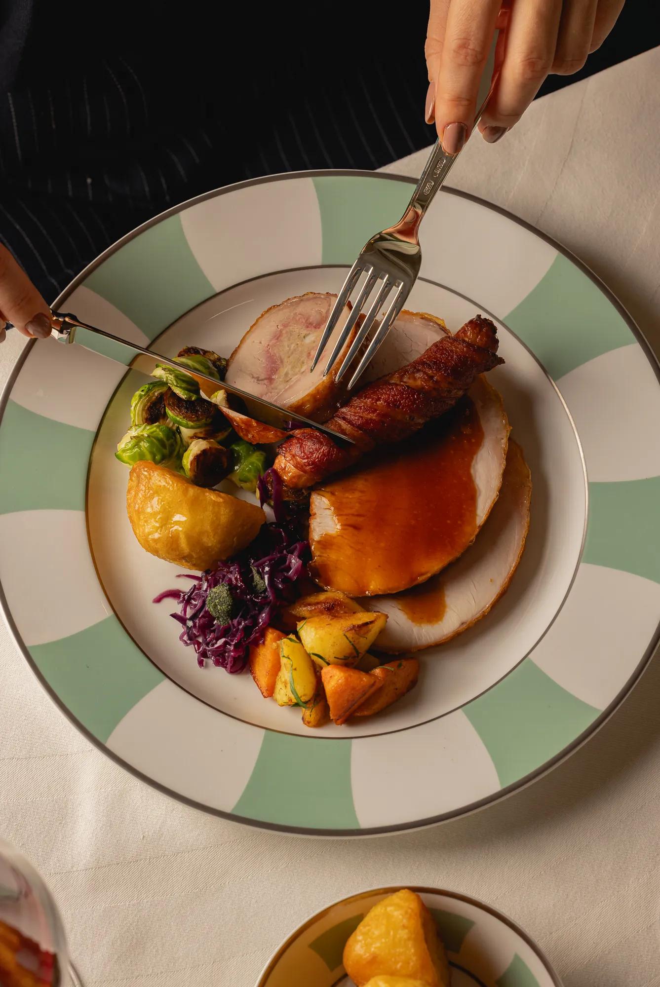 Roast turkey served with Brussels sprouts, red cabbage, roast potatoes and bacon-wrapped sausage, as a diner cuts into the meal with a knife and fork.