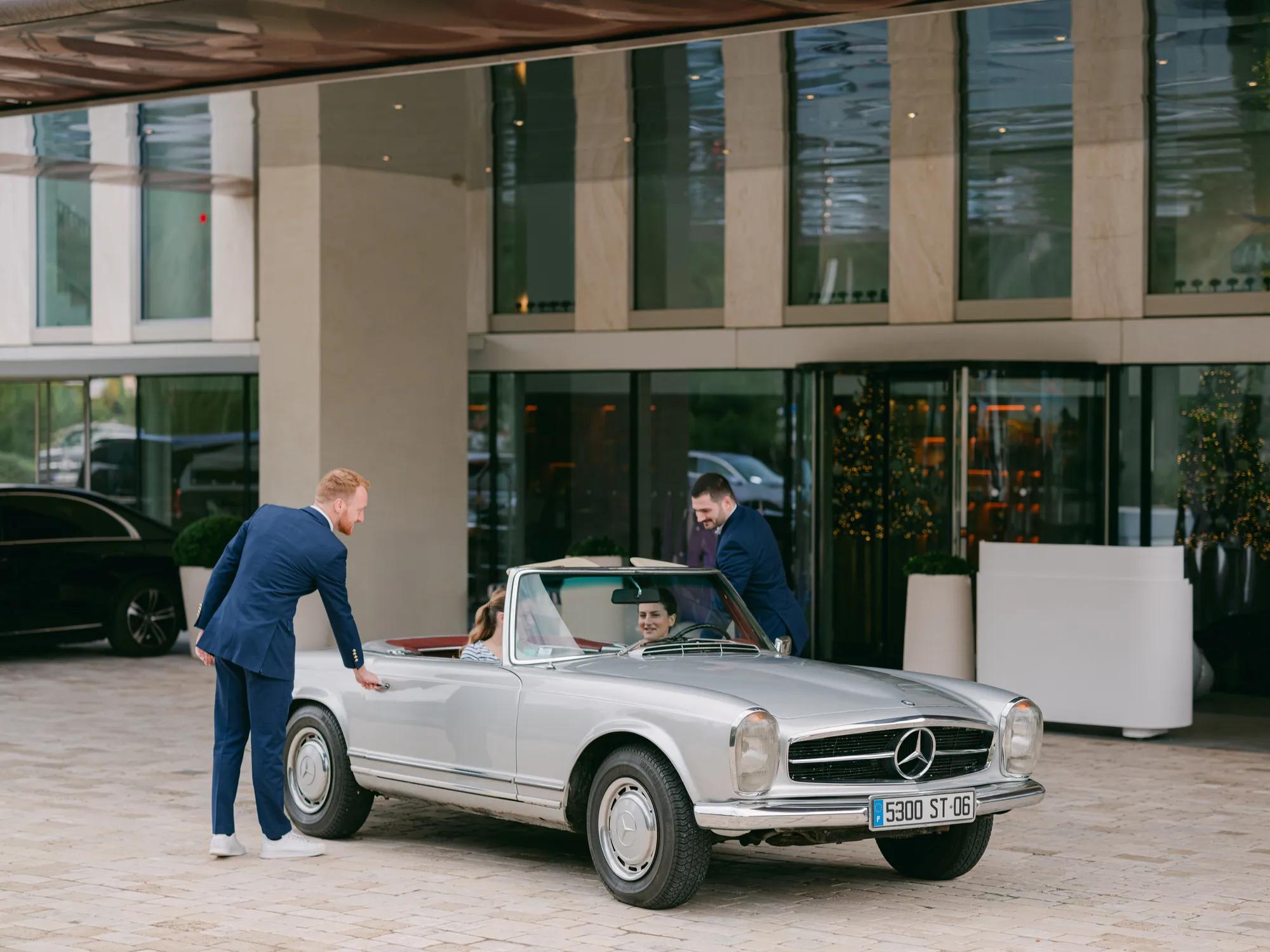 Valet staff welcome guests arriving in a silver vintage Mercedes convertible outside a modern hotel entrance.