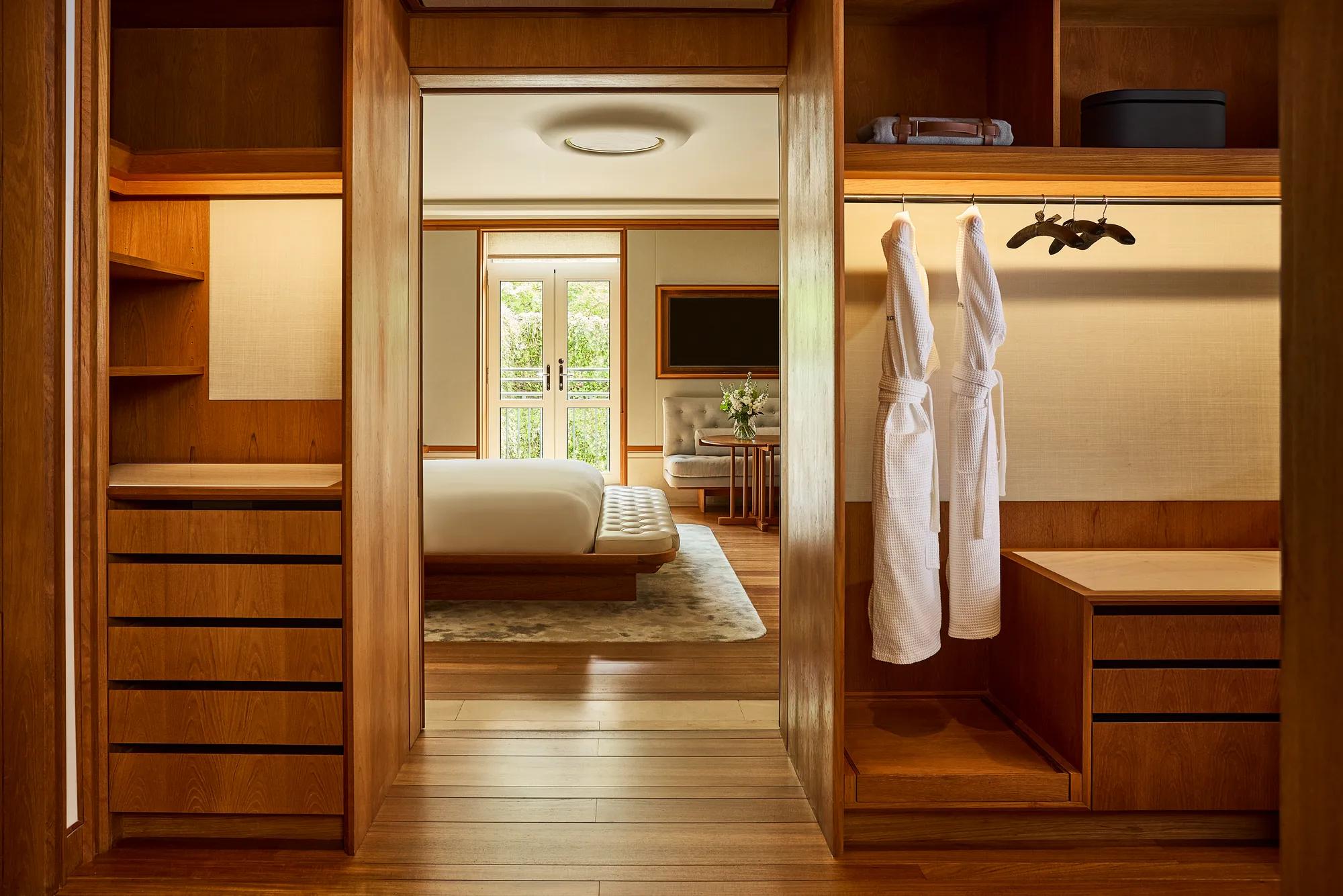 alk-in wardrobe with wooden cabinetry and two robes, opening onto a bedroom with balcony doors.