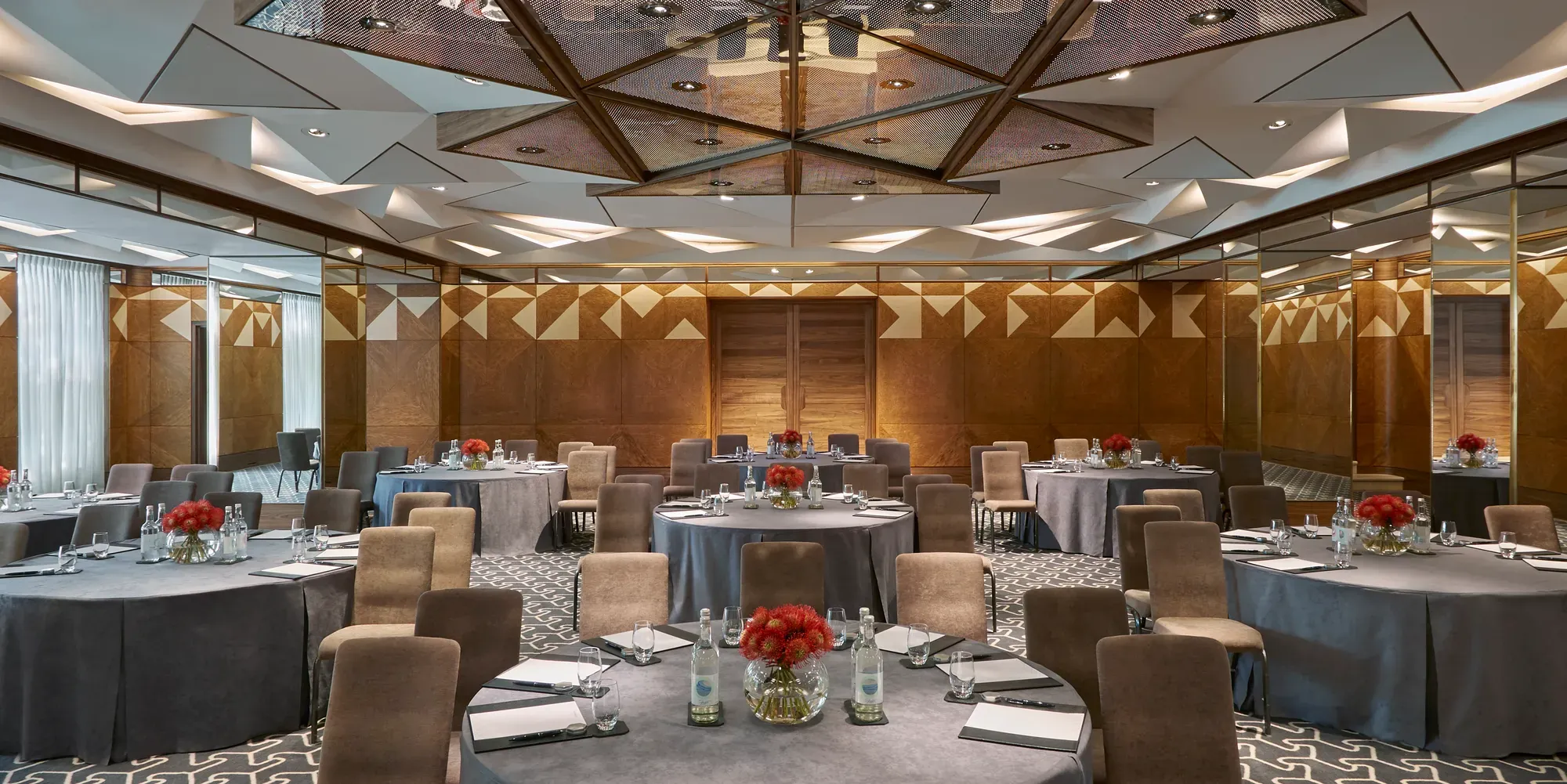 Conference room with round tables, grey tablecloths, notepads, water bottles, and red floral centerpieces under geometric mirrored ceiling.