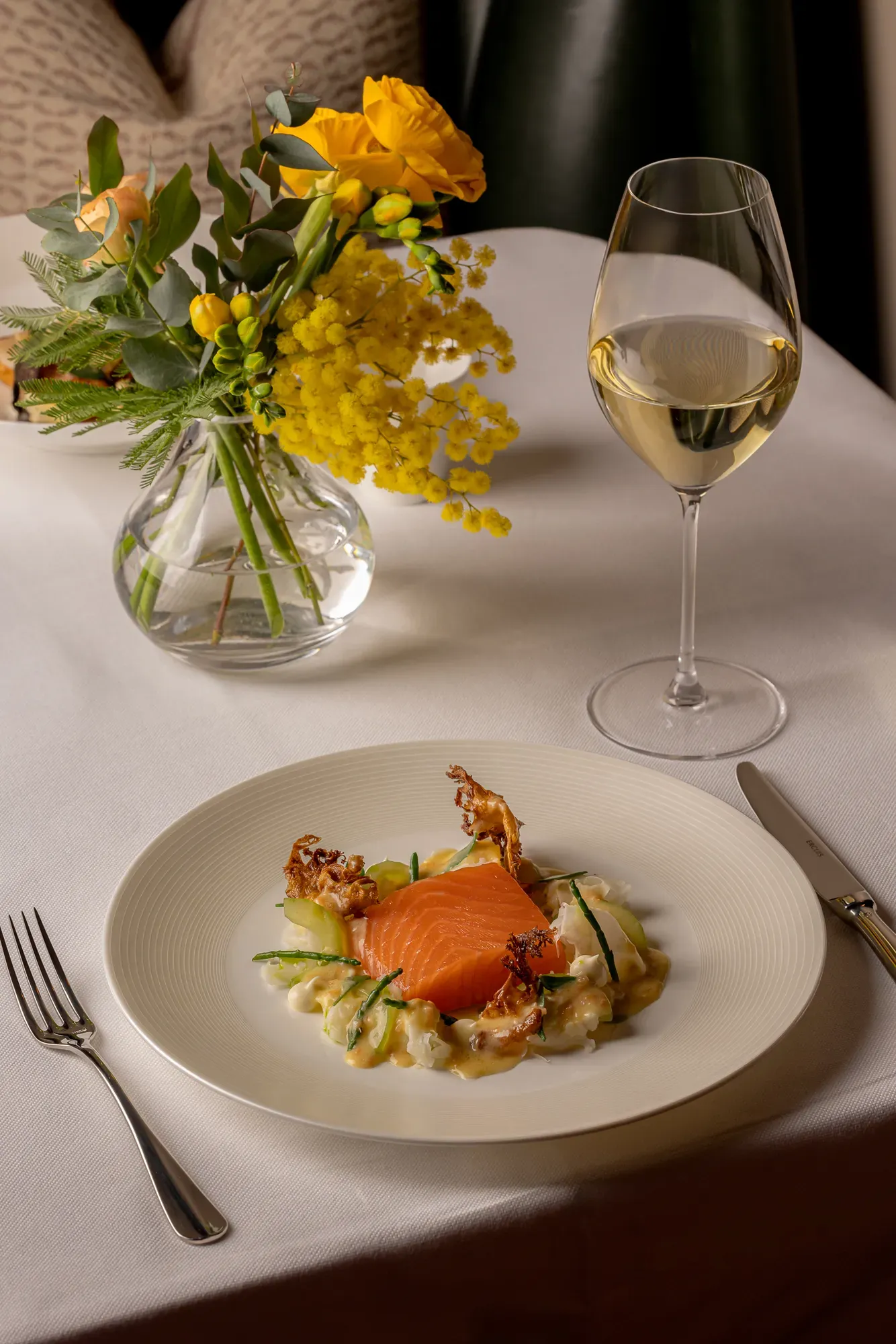 A fine dining dish of smoked salmon served on a creamy vegetable base with delicate garnishes, set on a white tablecloth beside a glass of white wine and a vase of yellow flowers.