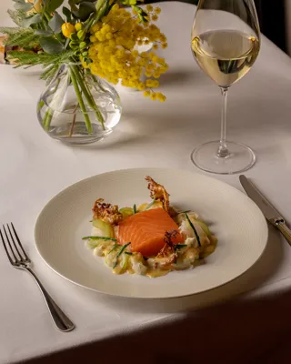A fine dining dish of smoked salmon served on a creamy vegetable base with delicate garnishes, set on a white tablecloth beside a glass of white wine and a vase of yellow flowers.