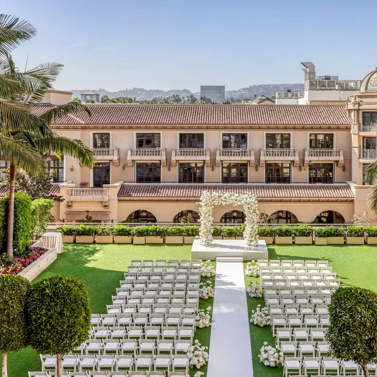 Garden Terrace wedding ceremony setup: long white aisle to a floral arch, rows of white chairs, palms and the domed rooftop beyond.