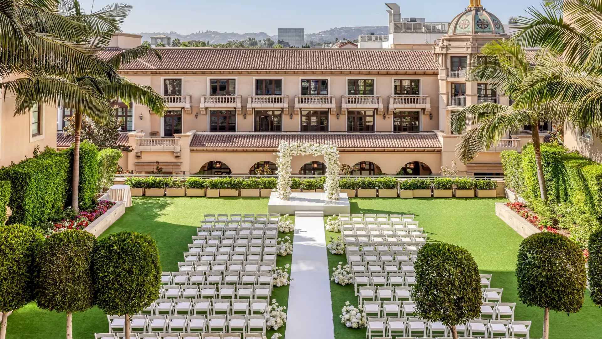 Garden Terrace wedding ceremony setup: long white aisle to a floral arch, rows of white chairs, palms and the domed rooftop beyond.