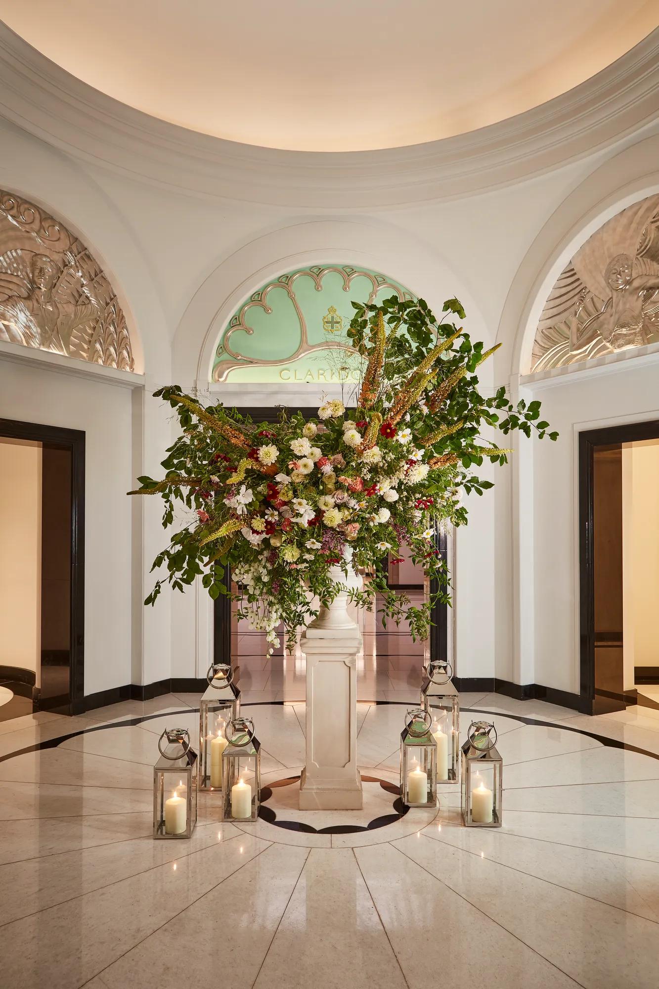 Elegant floral arrangement in the Claridge’s lobby, displayed on a marble pedestal surrounded by glass lanterns with candles, beneath an arched green glass panel and decorative plasterwork.