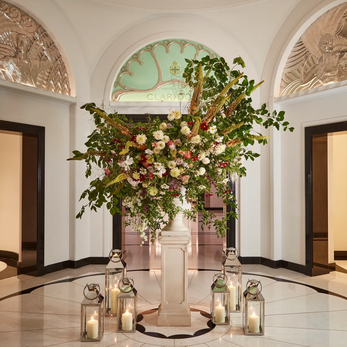 Elegant floral arrangement in the Claridge’s lobby, displayed on a marble pedestal surrounded by glass lanterns with candles, beneath an arched green glass panel and decorative plasterwork.