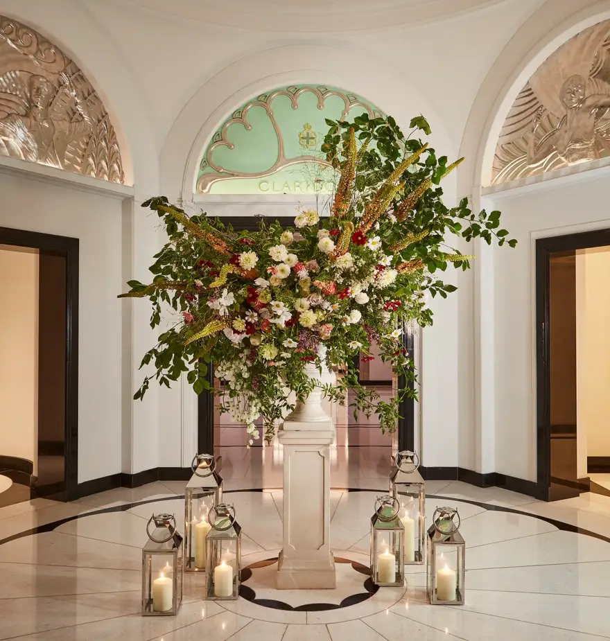 Elegant floral arrangement in the Claridge’s lobby, displayed on a marble pedestal surrounded by glass lanterns with candles, beneath an arched green glass panel and decorative plasterwork.