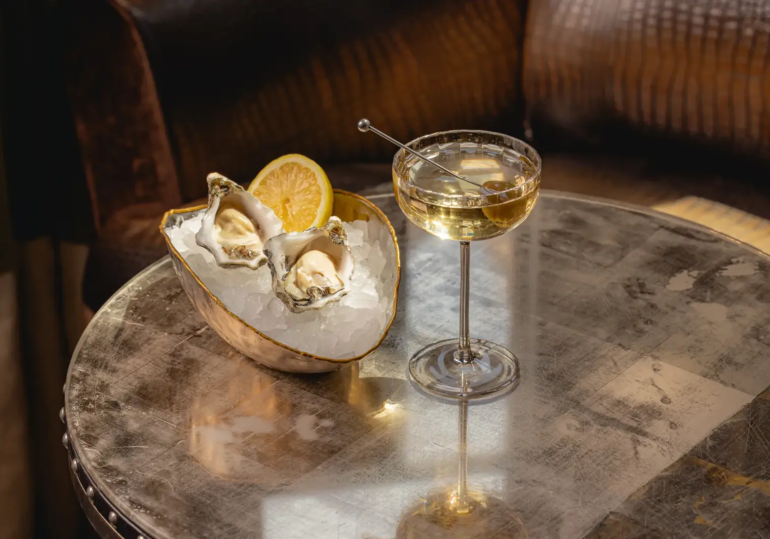 A close-up of a cocktail served in a coupe glass beside a bowl of oysters on ice with a lemon wedge, set on a metallic table in a softly lit, elegant bar setting.