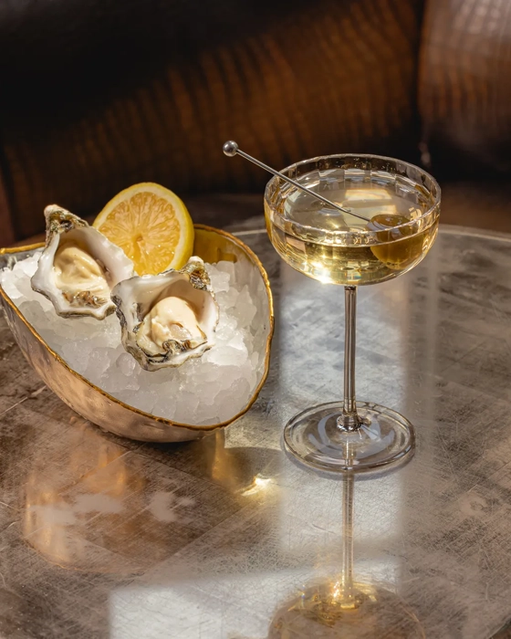 A close-up of a cocktail served in a coupe glass beside a bowl of oysters on ice with a lemon wedge, set on a metallic table in a softly lit, elegant bar setting.