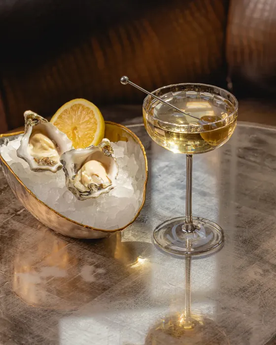 A close-up of a cocktail served in a coupe glass beside a bowl of oysters on ice with a lemon wedge, set on a metallic table in a softly lit, elegant bar setting.