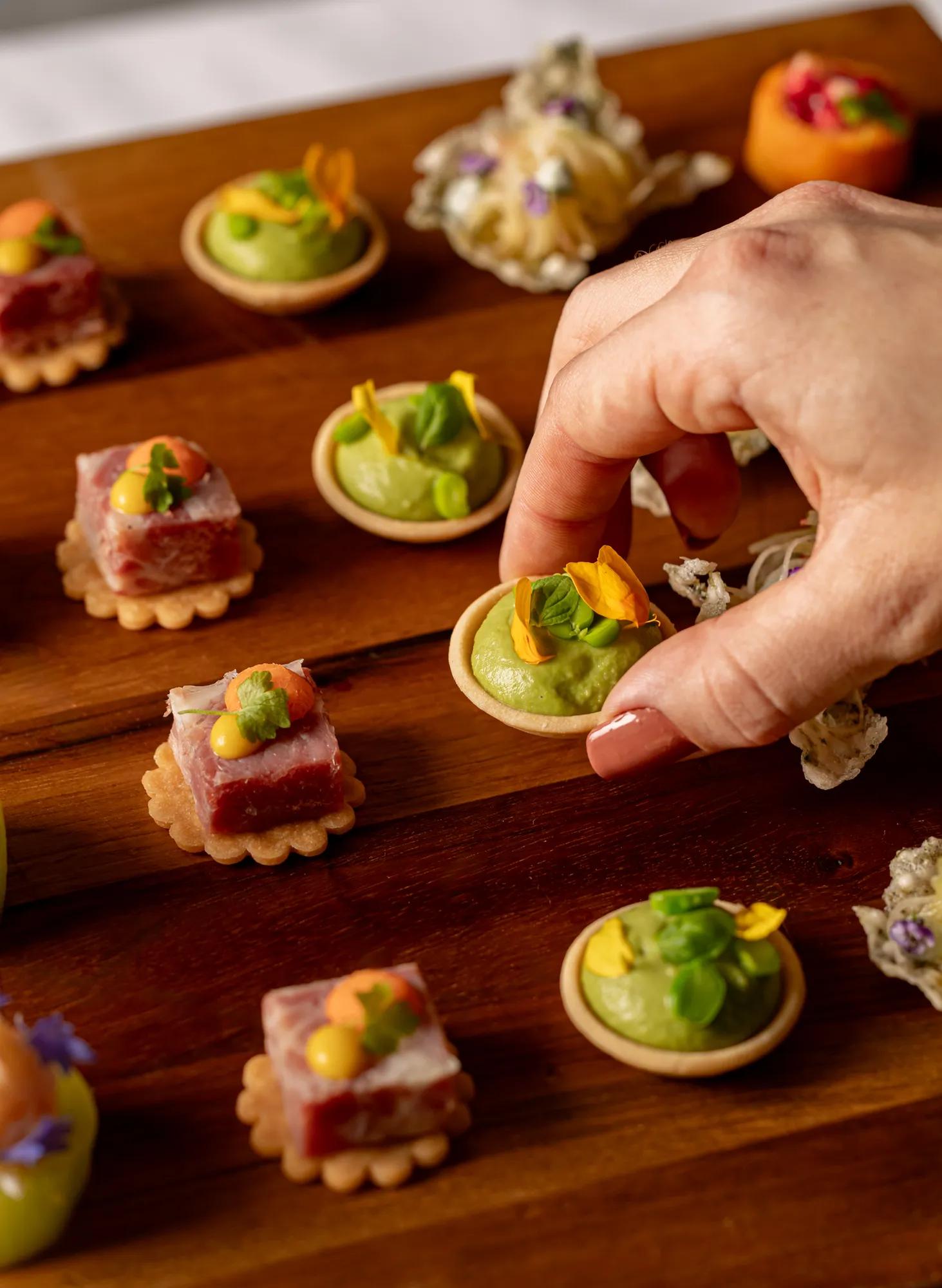 Hand selecting a canapé topped with green mousse, herbs and edible flowers from a wooden serving board.